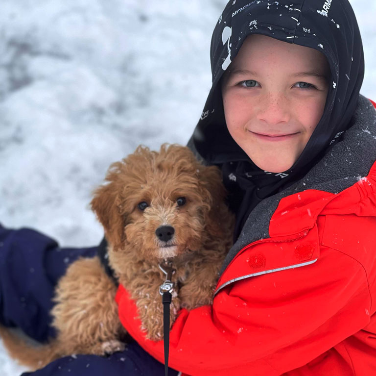 Litlle boy with red jacket holding a brown Cavapoo puppy while sitting in the snow