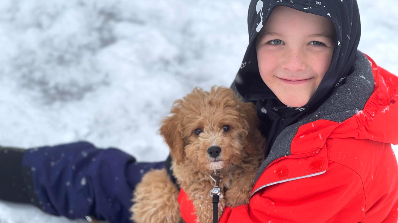 Little boy with red jacket holding a Cavapoo puppy and sitting in the snow