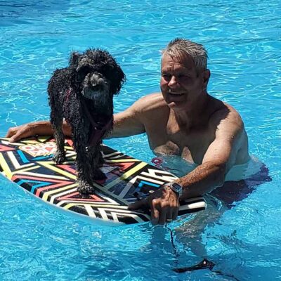 Man standing in a pool next to a black Cavapoo floating on top of a board
