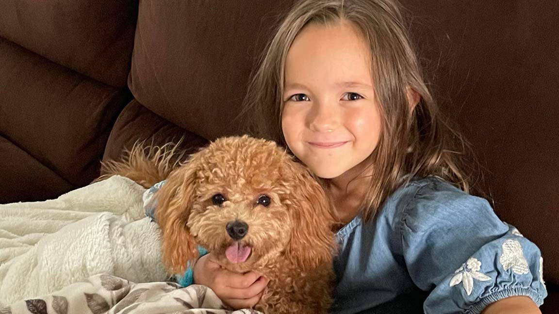 A young girl with long brown hair smiles while sitting on a couch, cuddling her small, fluffy brown Cavapoo dog.