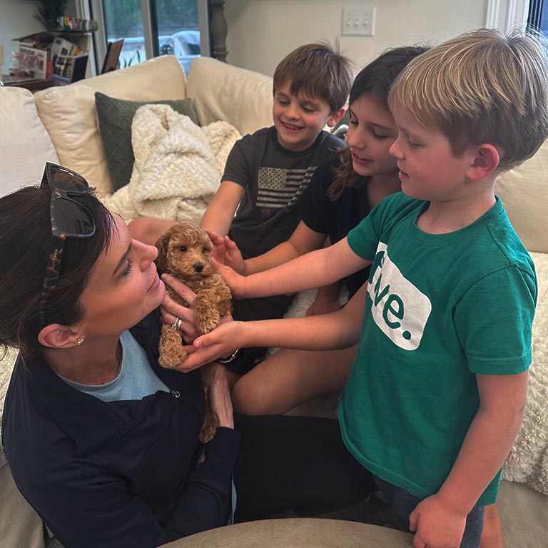 A woman and three children sit on a couch, all smiling and gently petting a small brown puppy.