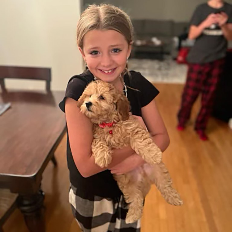A smiling young girl in black and plaid pajama pants holds a fluffy tan puppy indoors on a wooden floor.