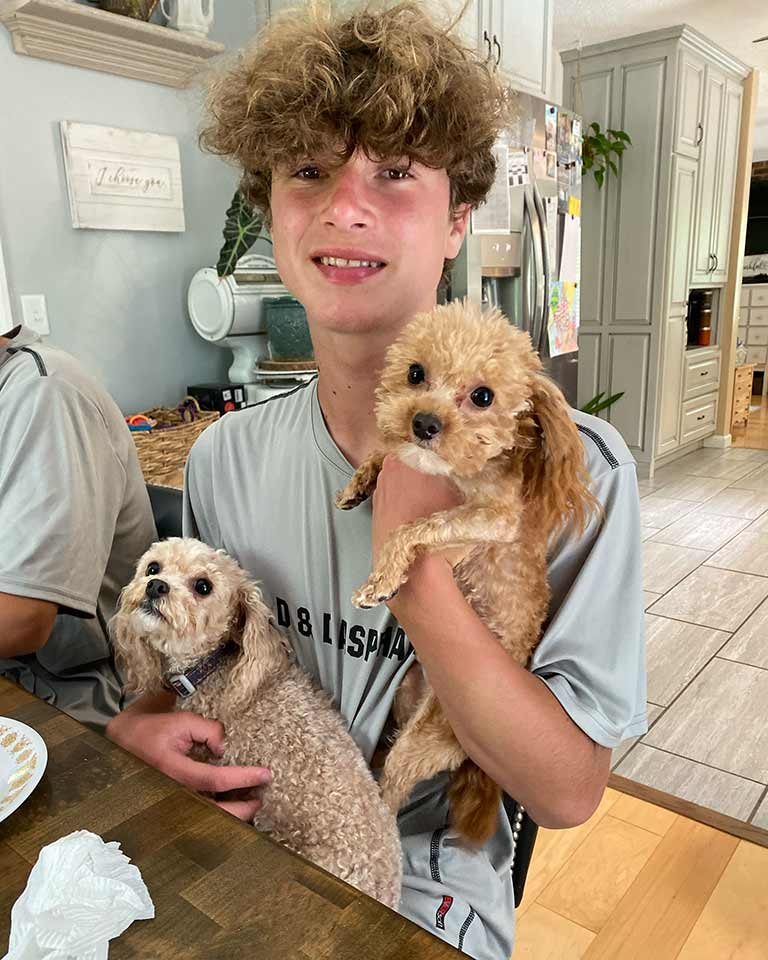 A teenage boy with curly hair sits at a kitchen table, smiling and holding two small, curly-haired dogs—one in his arm and the other on his lap. 