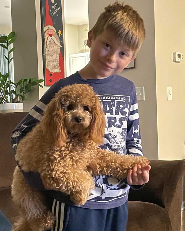 A young boy with light brown hair, wearing a blue Star Wars shirt, smiles while holding a small, curly-haired brown dog.