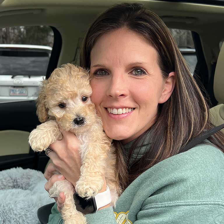 A smiling woman with long brown hair sits in a car, holding a small, curly-haired beige puppy close to her face.
