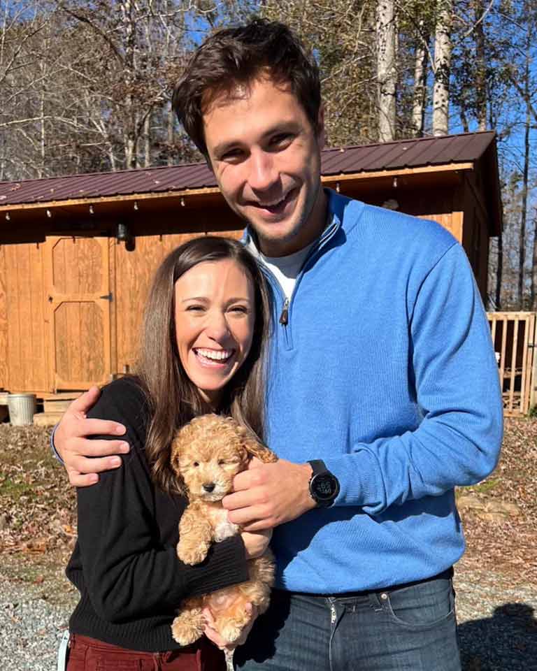 A smiling couple stands outdoors in front of a wooden shed, holding a small, fluffy tan puppy. The woman, in a black top, holds their newest puppy-proof companion while the man, in a blue sweater, has his arm around her. Trees are visible behind them.