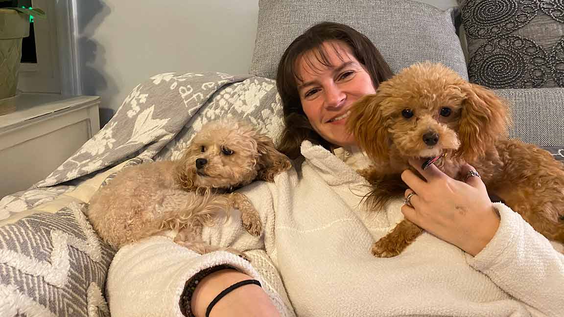 A woman in a white sweater is sitting on a puppy-proof couch, smiling and cuddling two Cavapoo dogs—one lying beside her and one in her arms. The couch features patterned cushions and a cozy blanket.
