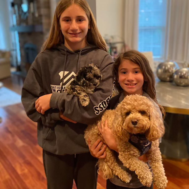 Two girls are smiling indoors, enjoying apartment living with a puppy. The older girl in a gray hoodie holds a small black and gray dog, while the younger in a dark shirt cuddles a light brown curly-haired pup.