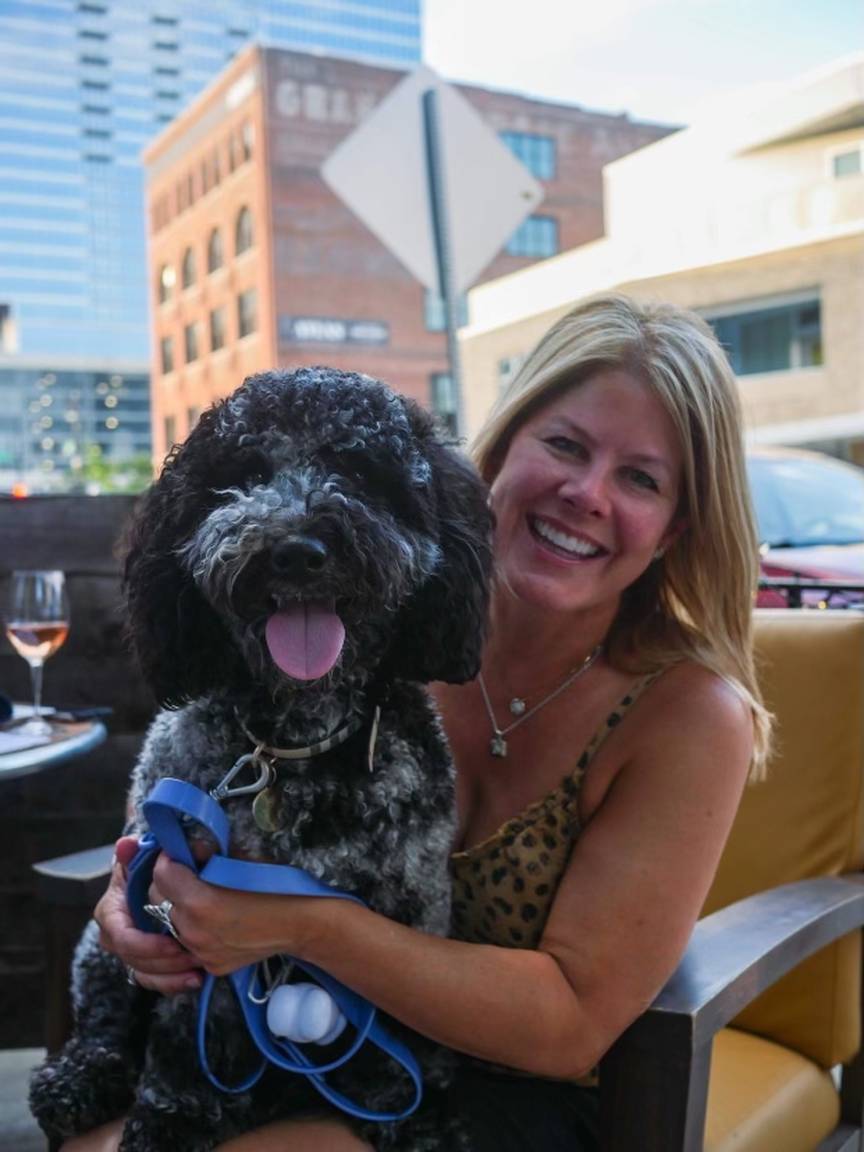 A smiling woman sits outdoors at a restaurant, happily hugging a large black and white curly-haired dog in her lap—enjoying quality time and mental stimulation for dogs. Buildings and a glass of wine are visible in the background.